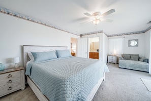 A bedroom with a bed, a nightstand, a dresser, a ceiling fan, and a door leading to another room. - Saltwater Revival - Stay with Seaside Vacations! (Chincoteague Island)