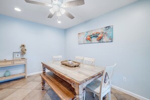 Comedor con una mesa de madera, sillas blancas, ventilador de techo y un cuadro en la pared.