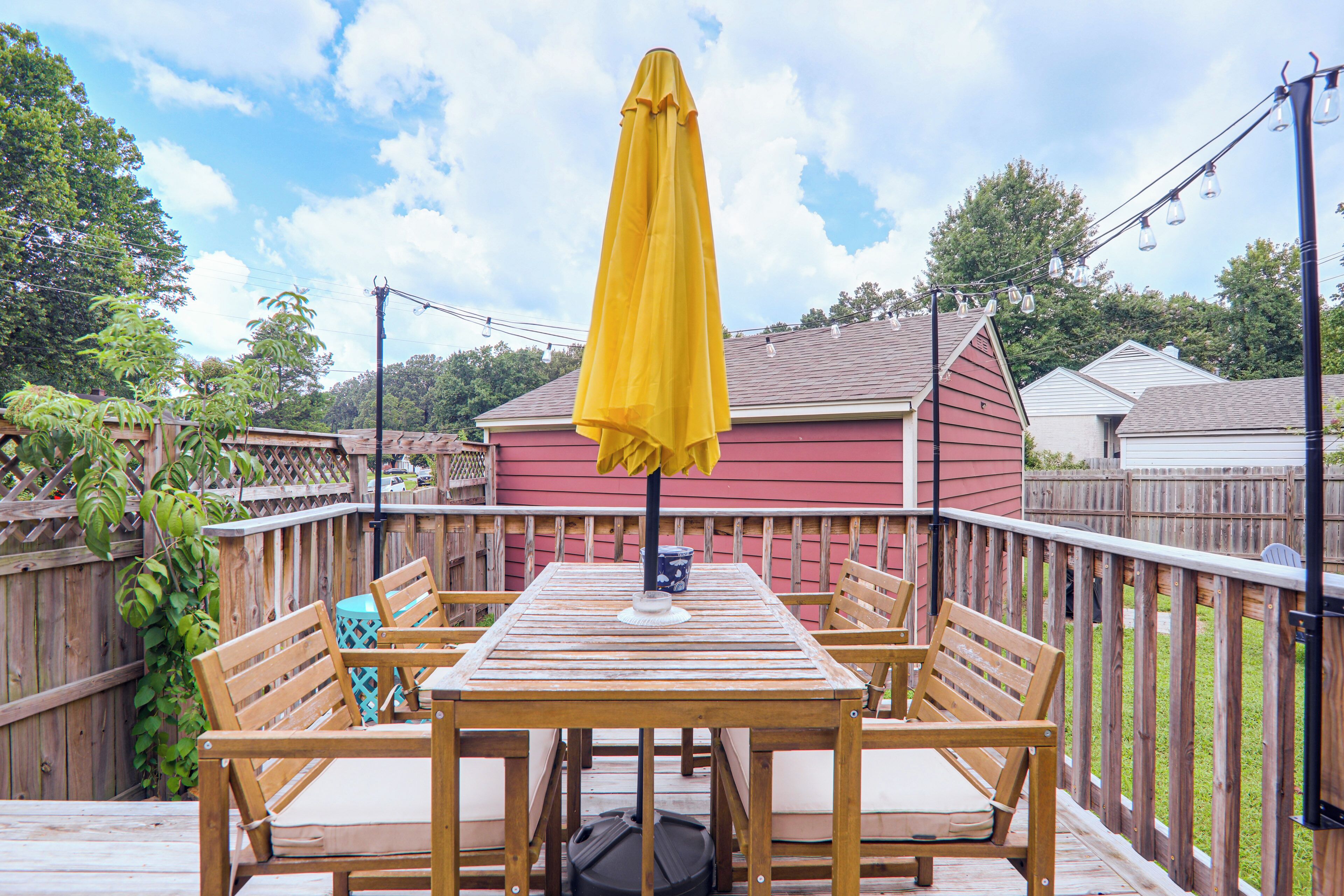 A wooden deck with a table and chairs, a yellow umbrella, and a red building in the background.