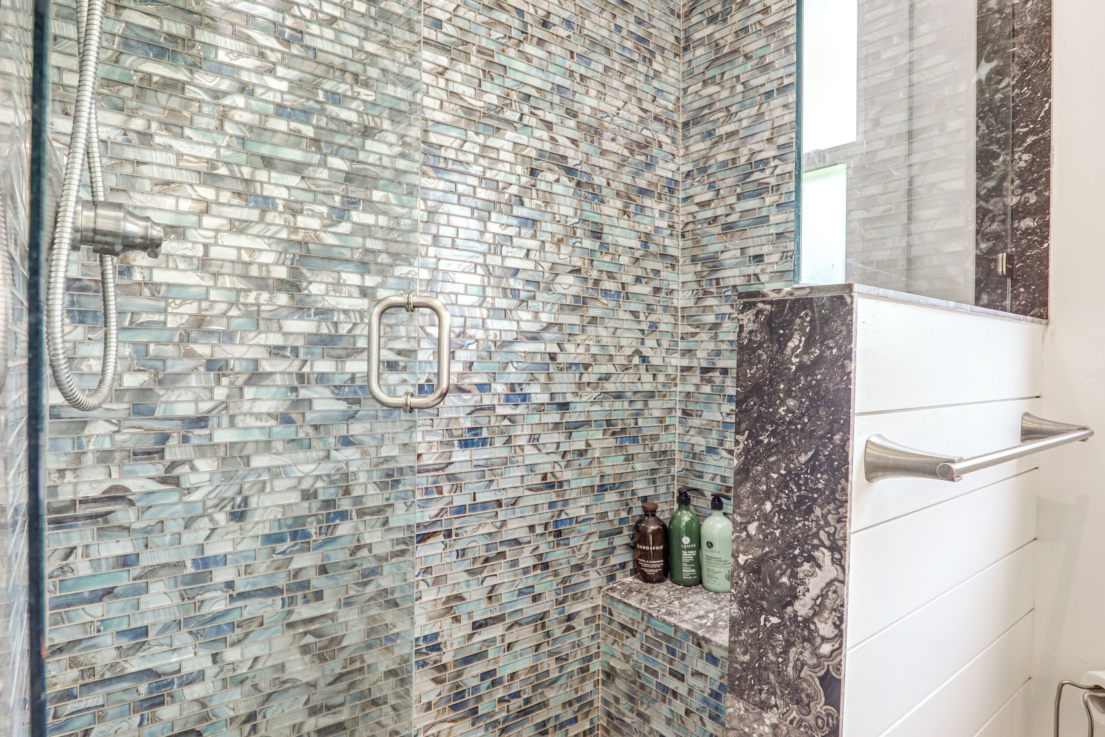 A bathroom with a mosaic tile wall, a shower with a handheld showerhead, and a shelf with toiletries.