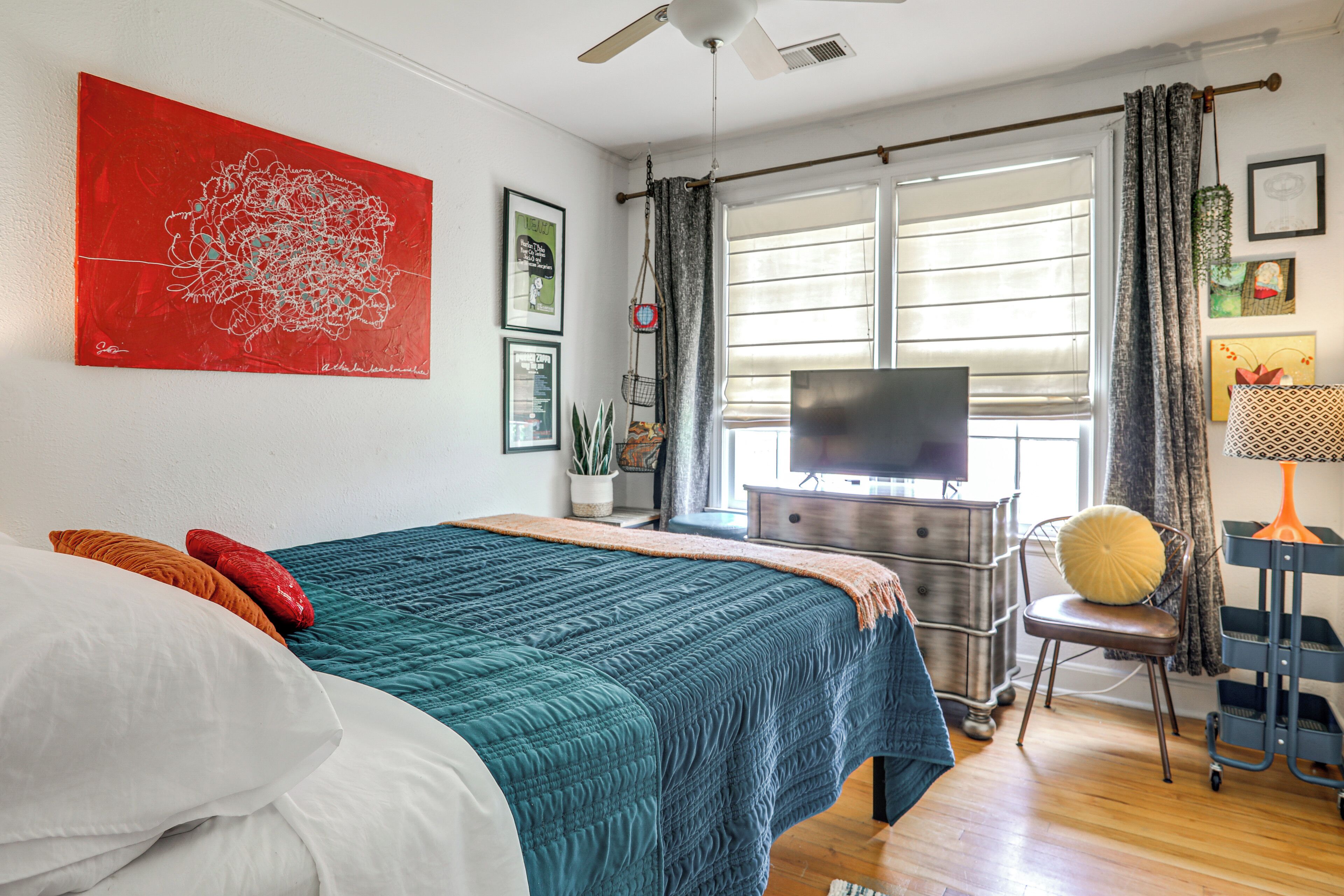 A bedroom with a bed, a TV, a window with curtains, and a red abstract painting on the wall.