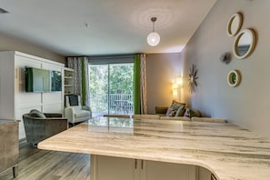 A modern kitchen with a marble countertop, a dining area with a table and chairs, a sofa, and a view of greenery through the window. - Near Grand Boulevard & Beaches! Coastal Condo (Miramar Beach)