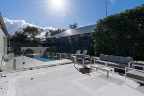 Ein Poolbereich mit Glasgalerie, Sitzgelegenheiten im Freien und Blick auf ein Haus im Hintergrund.