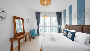 Ett hotellrum med en stor säng, ett träbord vid sängen, en blå stol, ett stort fönster med gardiner och en dekorativ väggdekoration.