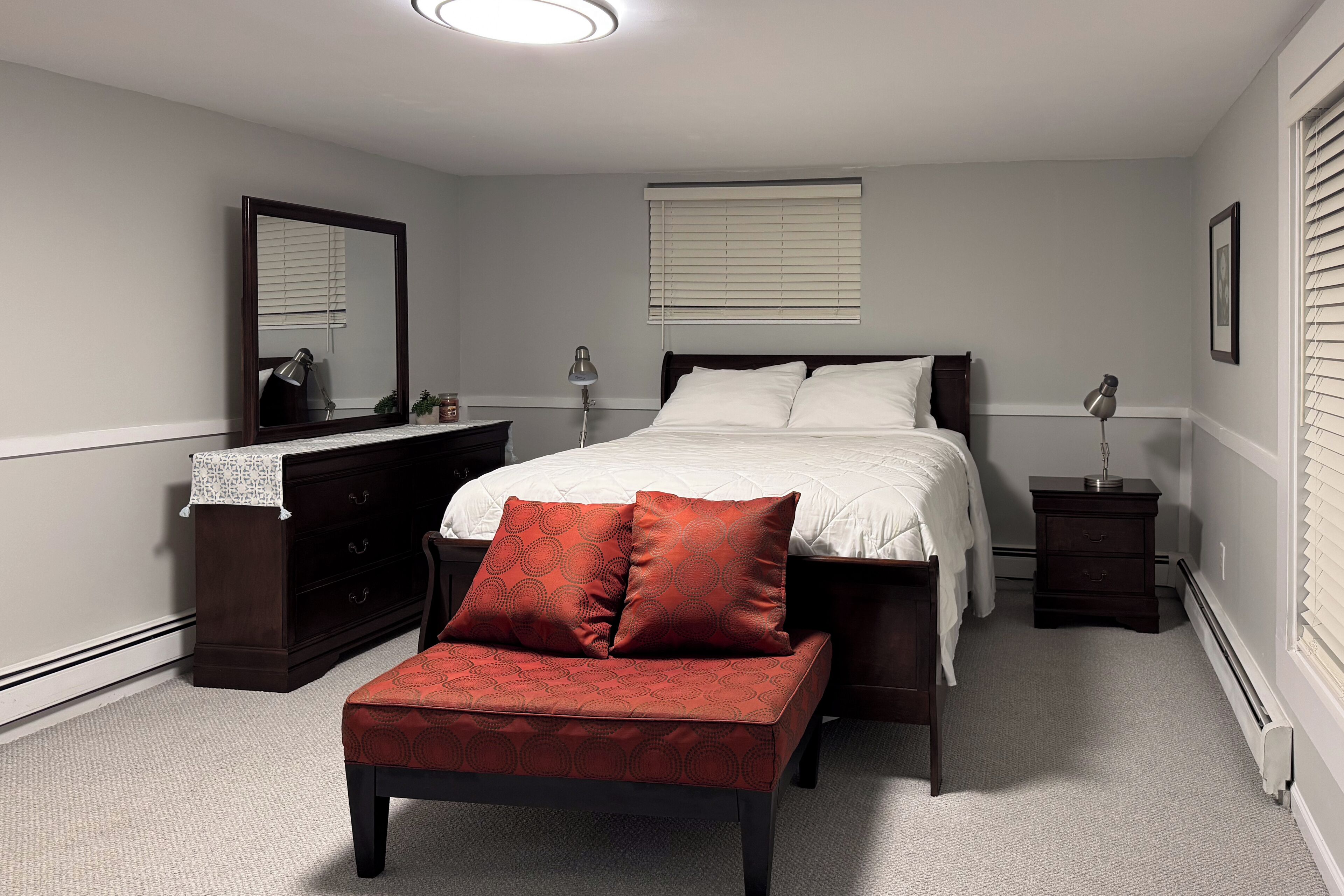 A bedroom with a bed, a dresser, a bench, a bedside table, a lamp, a mirror, and a window with blinds.