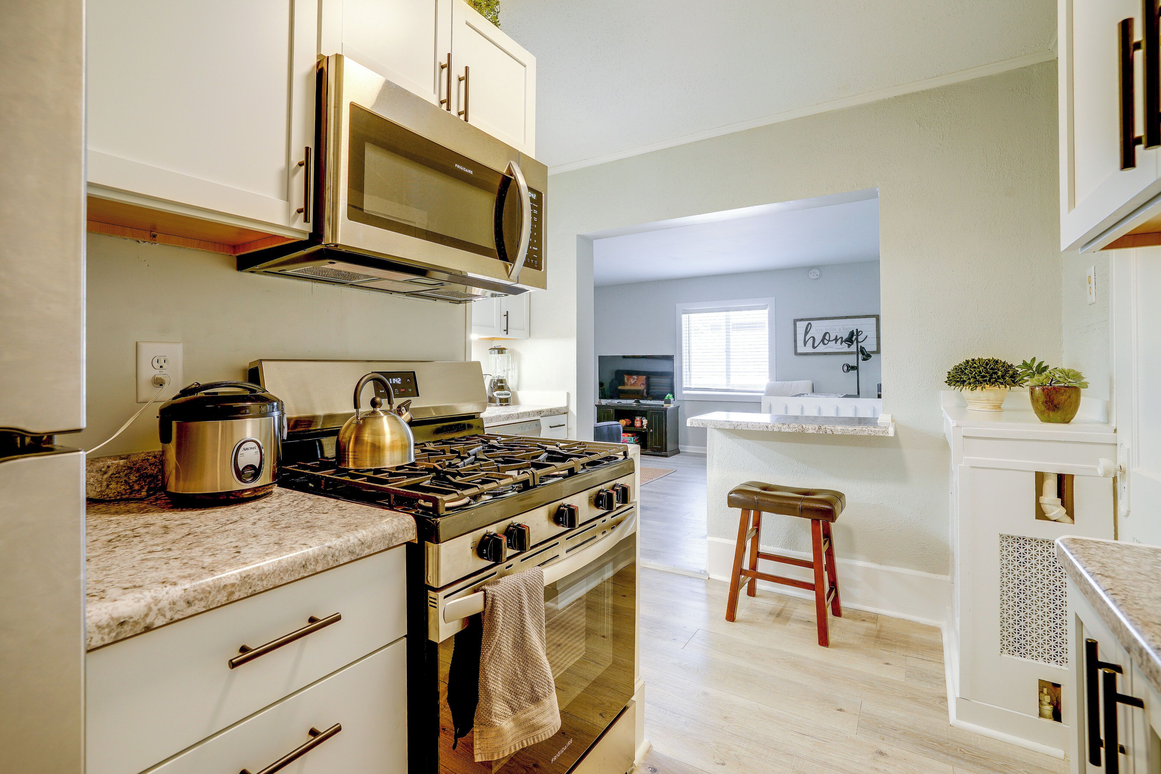 A kitchen with a stove, oven, and microwave. There is a wooden stool and a countertop with a kettle.