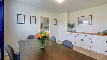 A dining area with a wooden table, chairs, a vase with flowers, framed pictures on the wall, and a kitchen area with cabinets and a countertop.
