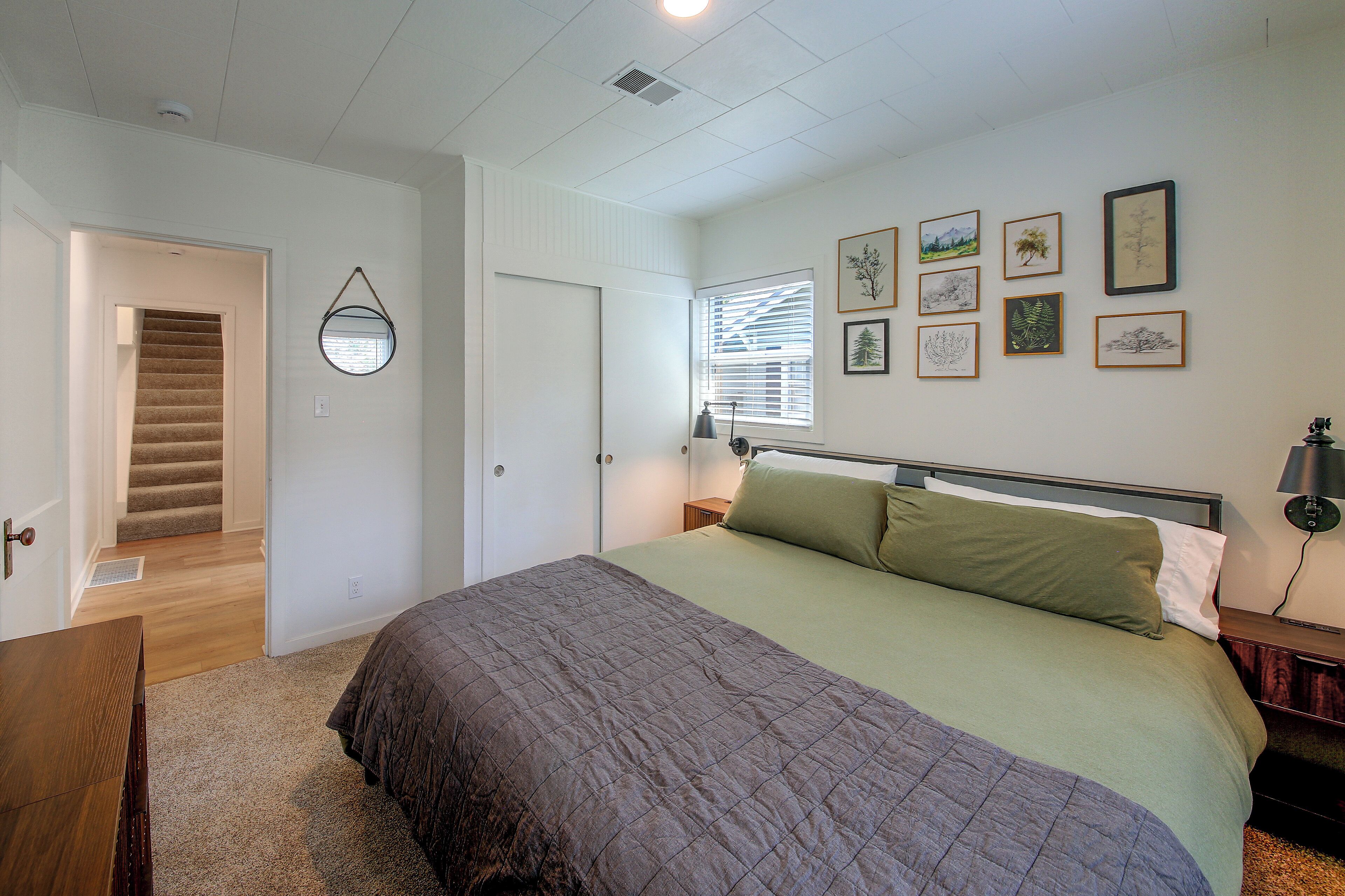 A bedroom with a bed, a nightstand, a lamp, a window with blinds, a door, and a staircase leading to another room.