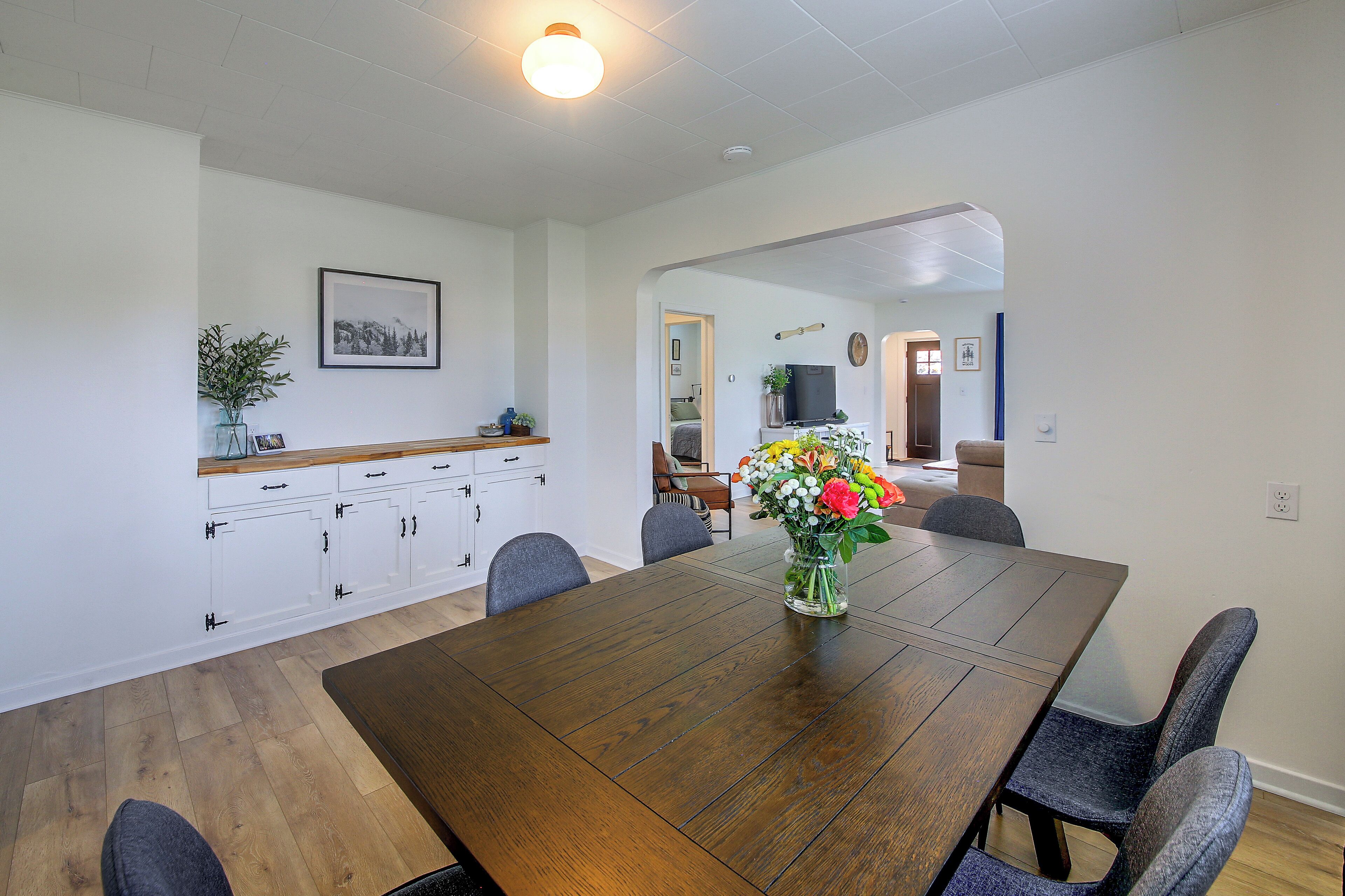 A dining area with a wooden table, chairs, a vase with flowers, and a kitchen area with cabinets and a countertop.