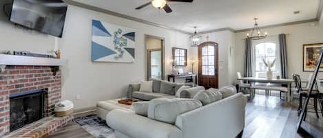 A living room with a fireplace, a grey sectional sofa, a ceiling fan, and a dining area with a table and chairs.