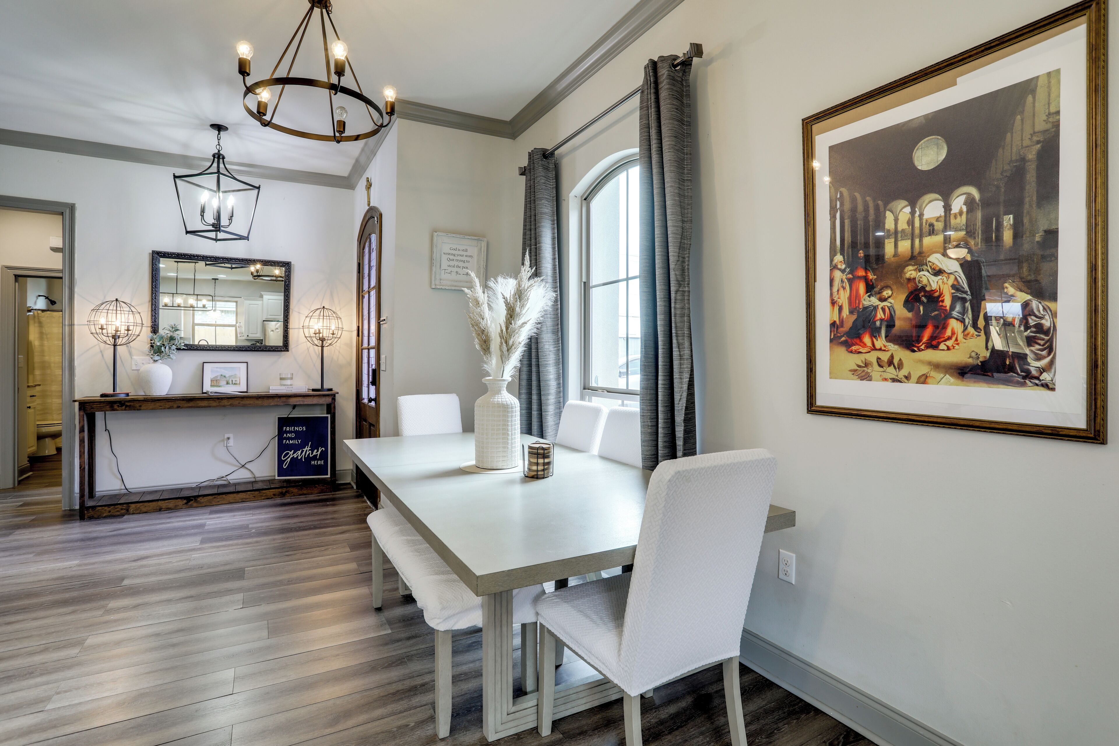 A dining area with a white table, chairs, and a chandelier. There is a large framed painting on the wall and a window with curtains.