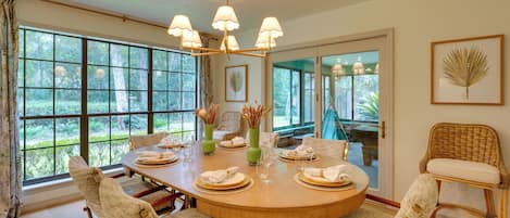A dining room with a round table set for six, surrounded by wicker chairs, large windows, and a chandelier.