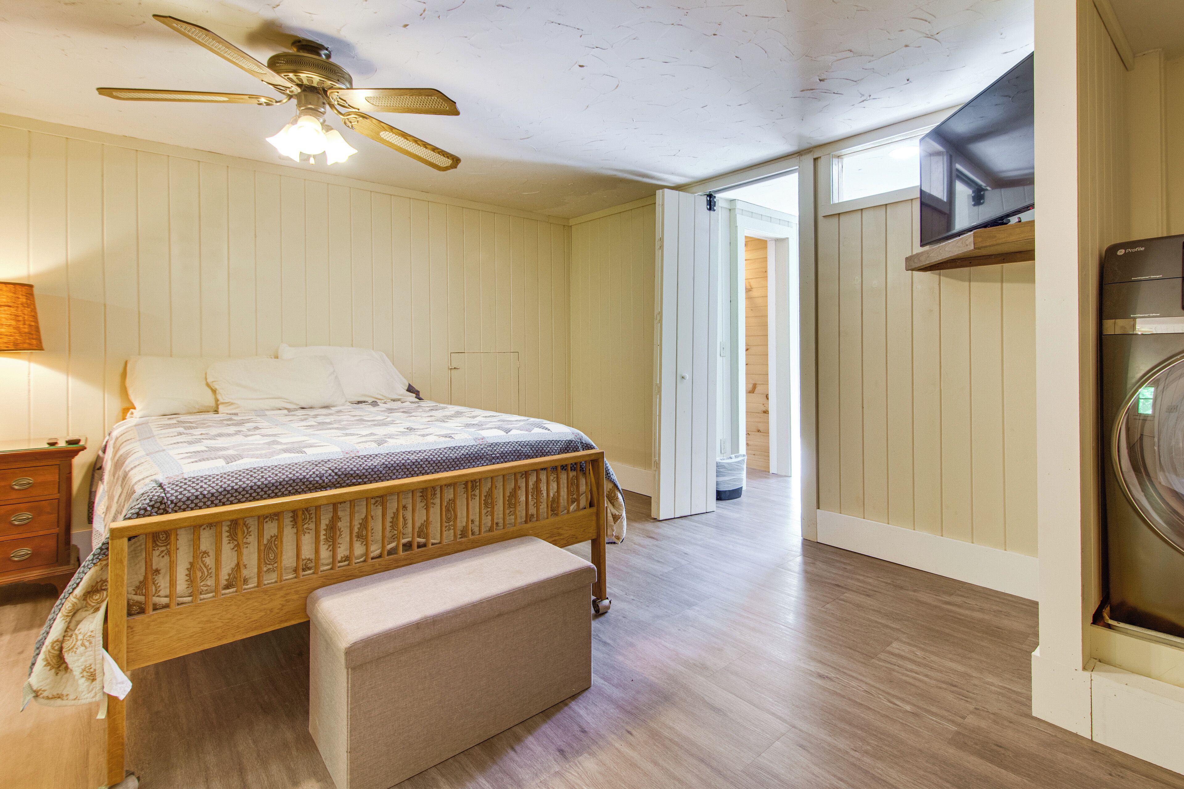 A bedroom with a bed, a wooden bench, a ceiling fan, a TV, and a washing machine.