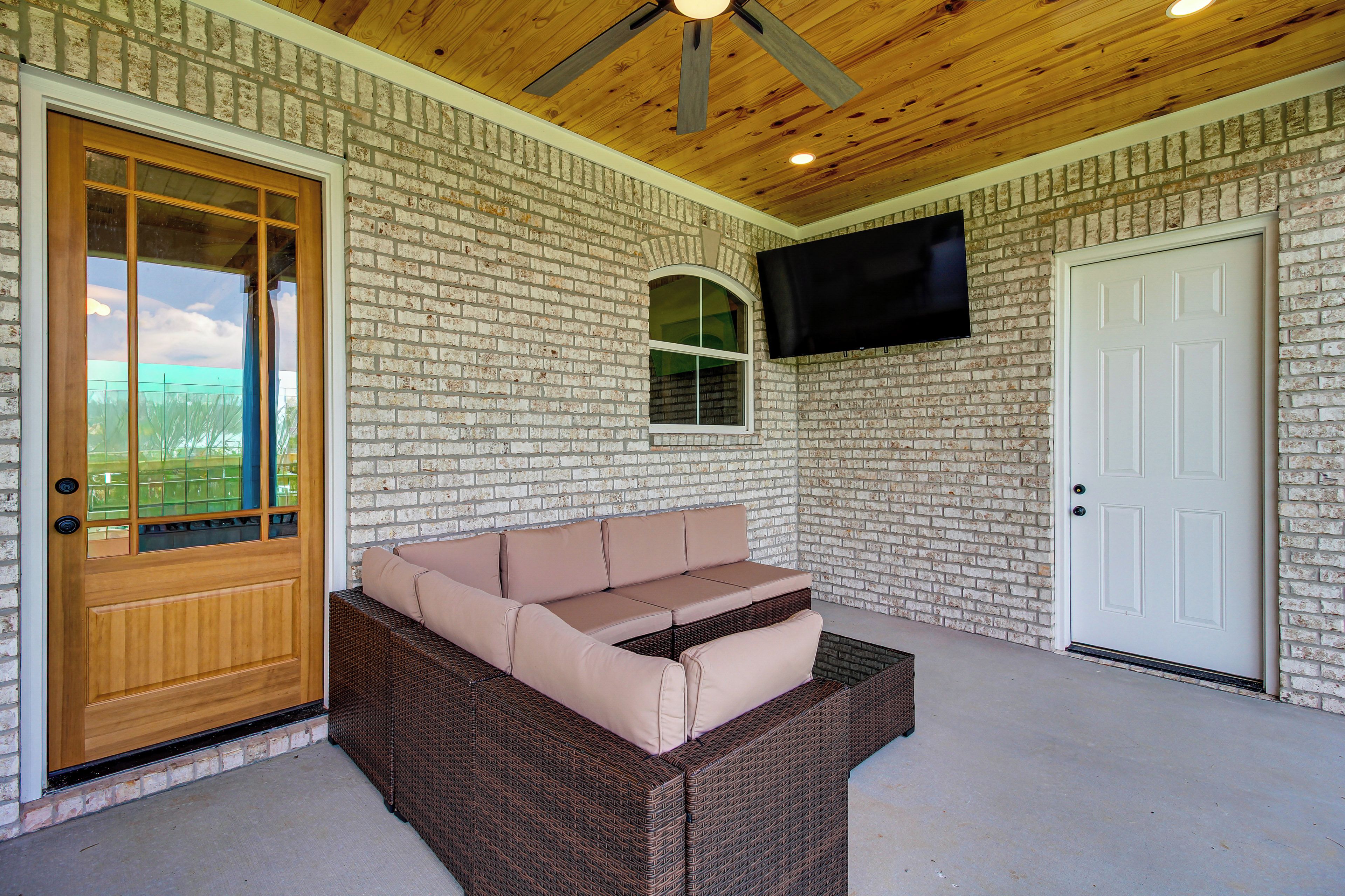 A covered porch with a wicker sofa, a wooden door, a brick wall, and a mounted TV.