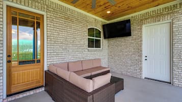 A covered porch with a wicker sofa, a wooden door, a brick wall, and a mounted TV.