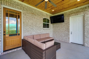 A covered porch with a wicker sofa, a wooden door, a brick wall, and a mounted TV.