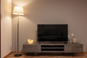 Ein modernes Wohnzimmer mit einem Flachbildfernseher auf einem grauen Schrank, einer Stehlampe und Kerzen auf einem kleinen Tisch.