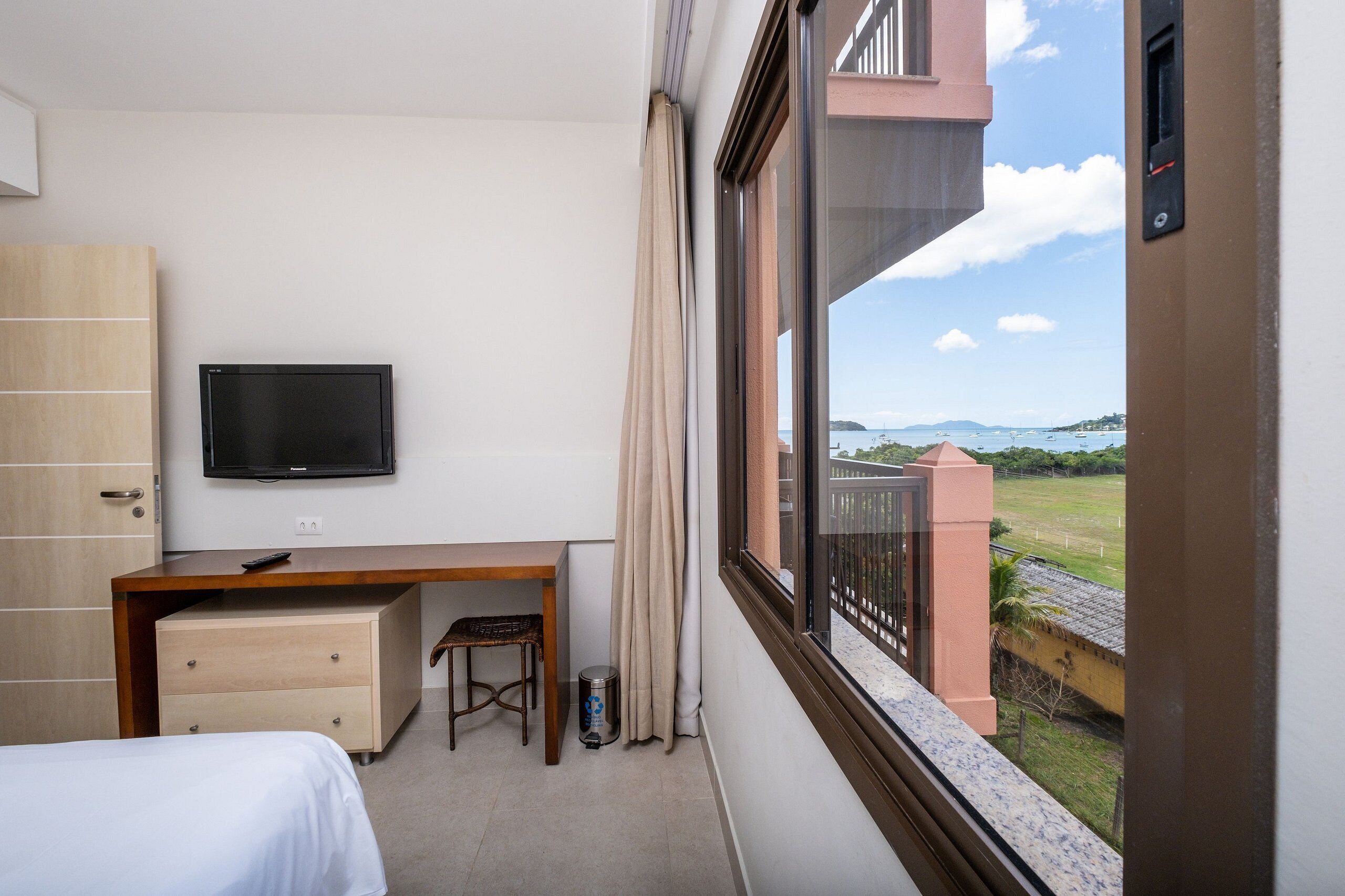 A hotel room with a bed, a desk, a television, and a window with a view of the sea and buildings.
