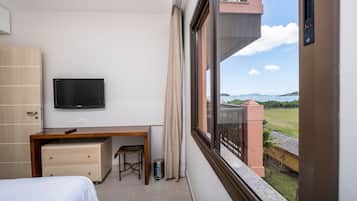A hotel room with a bed, a desk, a television, and a window with a view of the sea and buildings.