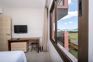 A hotel room with a bed, a desk, a television, and a window with a view of the sea and buildings. - Great ap With Pool in Resort in Jurere Jbv157 (Florianópolis)