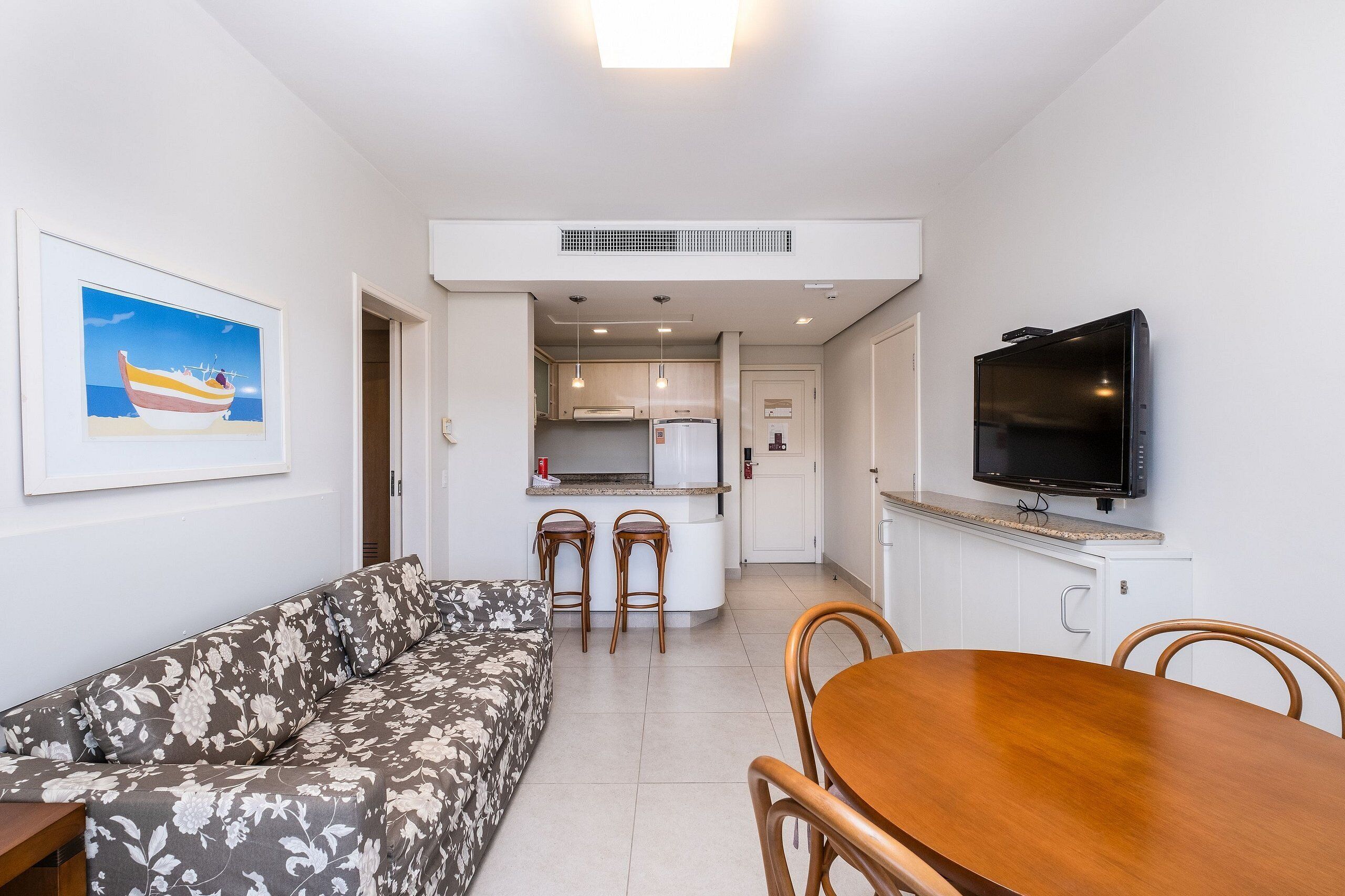 A living room with a sofa, a dining table with chairs, a kitchen area with a fridge, and a TV mounted on the wall.
