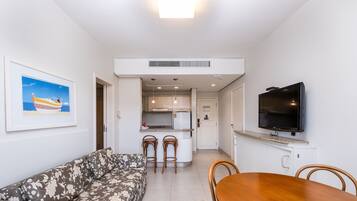 A living room with a sofa, a dining table with chairs, a kitchen area with a fridge, and a TV mounted on the wall.