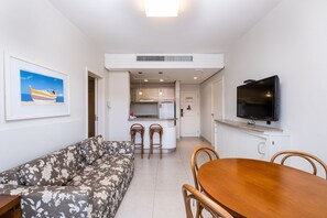 A living room with a sofa, a dining table with chairs, a kitchen area with a fridge, and a TV mounted on the wall. - Great ap With Pool in Resort in Jurere Jbv157 (Florianópolis)