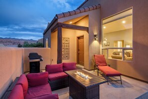 Un patio exterior con un pozo de fuego, asientos con cojines rojos y vistas al interior a través de una puerta de vidrio.