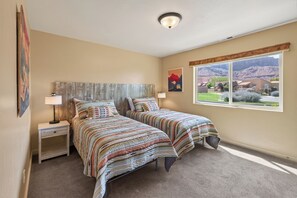 Une chambre avec deux lits, une table de chevet, une lampe, une fenêtre donnant sur les montagnes et un tableau au mur.