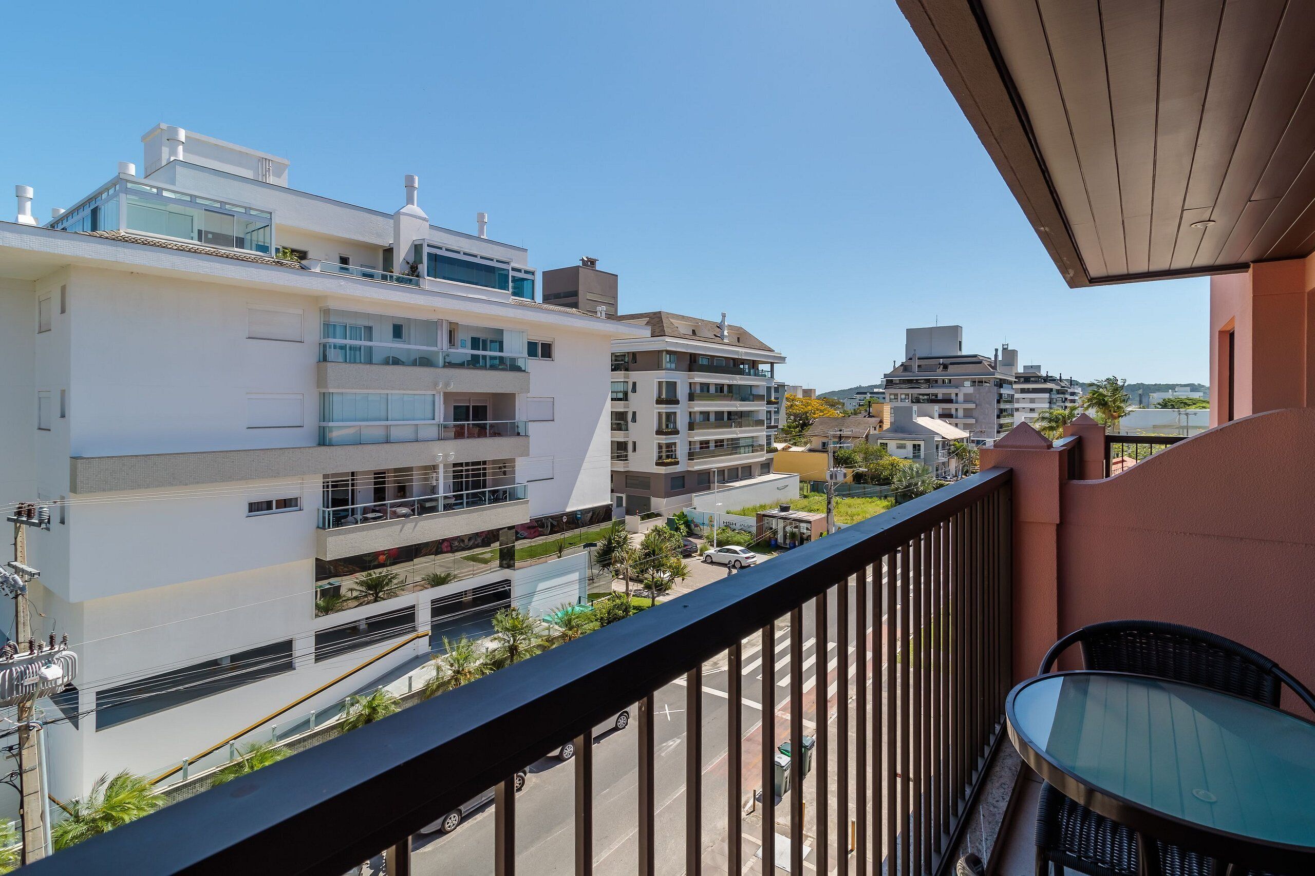 Varanda com mesa e cadeiras de vidro, vista para uma rua com edifícios e céu claro.