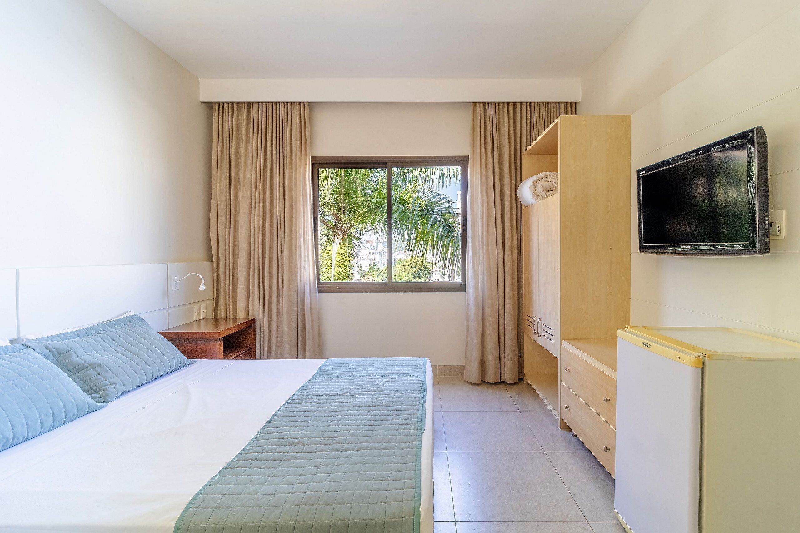 Quarto de hotel com cama, televisão, escrivaninha e janela com cortinas.