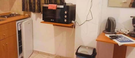 A compact room with a kitchenette, a television mounted on the wall, a small trash bin, and a desk with a chair.