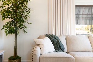 A living room with a sofa, a potted tree, and a window with curtains. - Clover Cottage - 1 mile to ND - Cozy Getaway (South Bend)