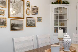 A dining area with white chairs, a wooden table, and a white cabinet with glass doors. - Clover Cottage - 1 mile to ND - Cozy Getaway (South Bend)