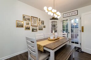 A dining area with a wooden table, white chairs, a chandelier, and framed pictures on the wall. - Clover Cottage - 1 mile to ND - Cozy Getaway (South Bend)