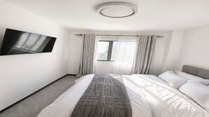 Ein Hotelzimmer mit Bett, Wandfernseher, einem Fenster mit Vorhängen und einer Deckenleuchte.