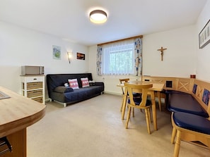 Ein Wohnzimmer mit einer Couch, einem Esstisch mit Stühlen, einem Fernseher auf einem Schrank und einem Fenster mit Vorhängen.
