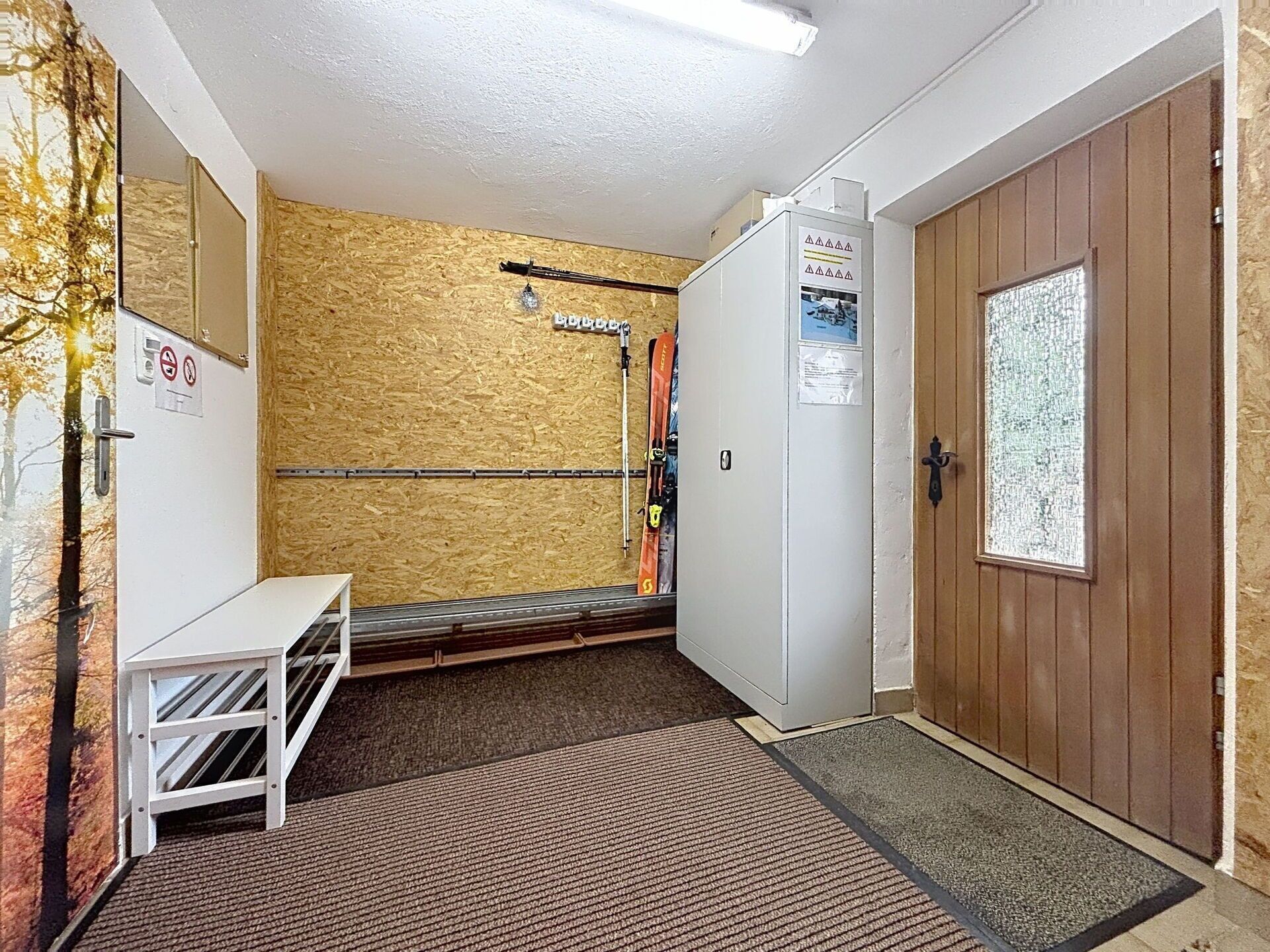 Une petite pièce fermée, dotée d’une porte en bois, d’un mur blanc avec une fenêtre et d’une étagère en métal.