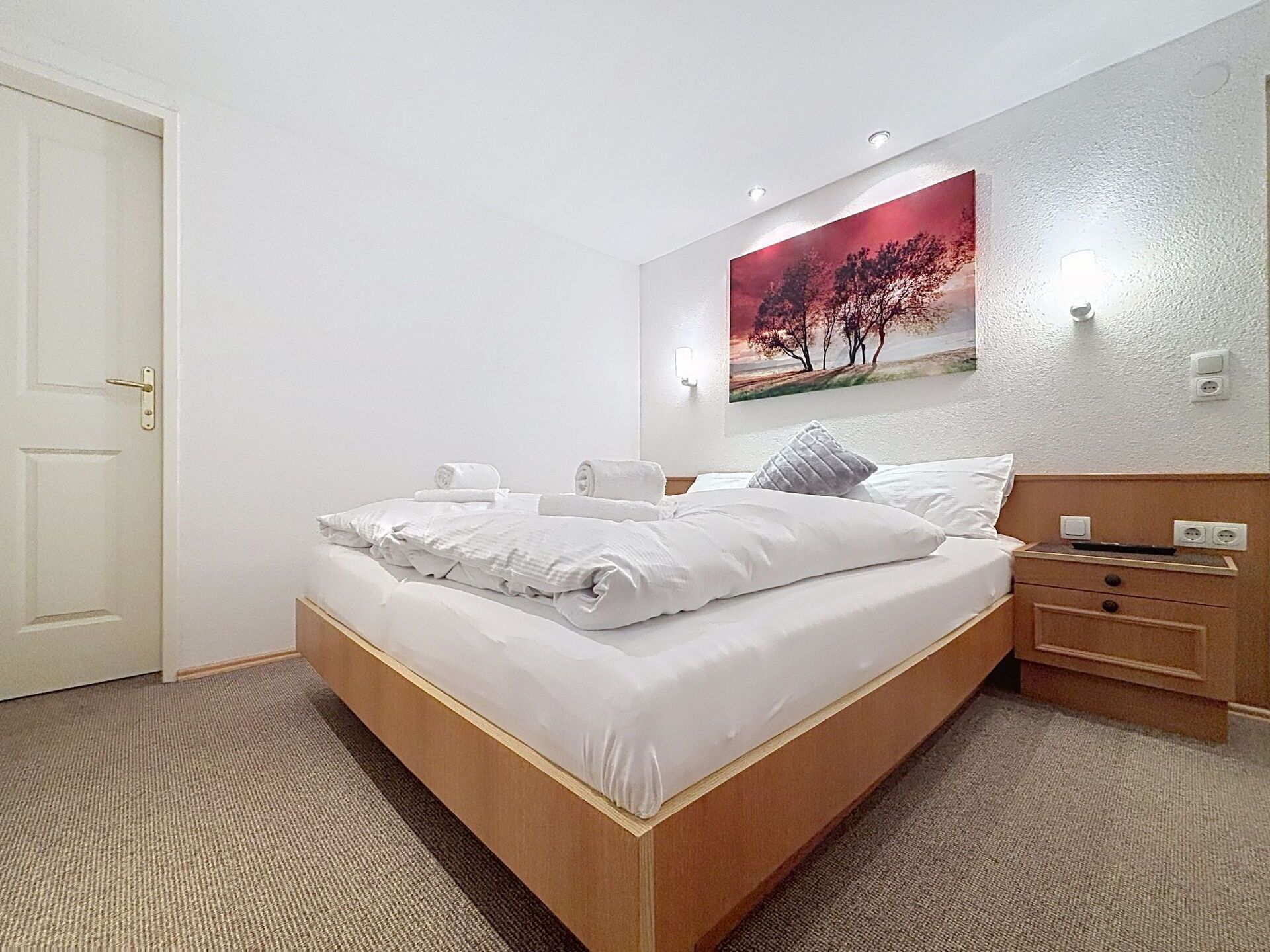 A hotel room with a bed, a nightstand, a wall-mounted lamp, and a painting.