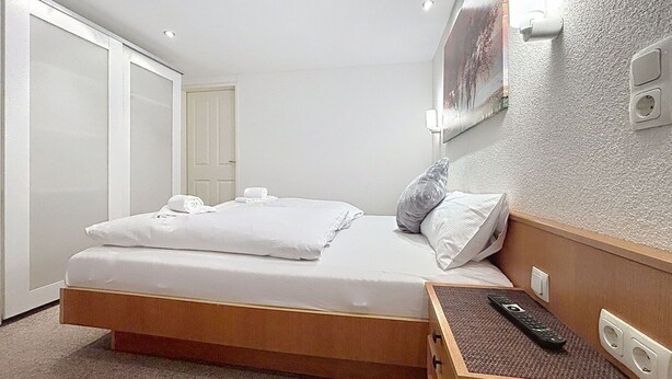 Quarto de hotel com cama, mesa de cabeceira com controle remoto, porta e luminária na parede.