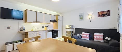 A compact living space with a dining table, kitchenette, sofa, and a flat-screen TV mounted on the wall.