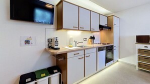 Uma cozinha compacta com forno embutido, micro-ondas e máquina de café. Há armários com puxadores e uma bancada com pia.