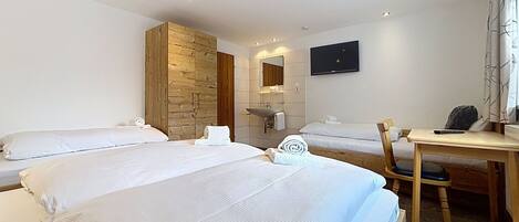 A hotel room with two beds, a wooden wardrobe, a bathroom with a mirror and sink, a desk with a chair, and a TV mounted on the wall.