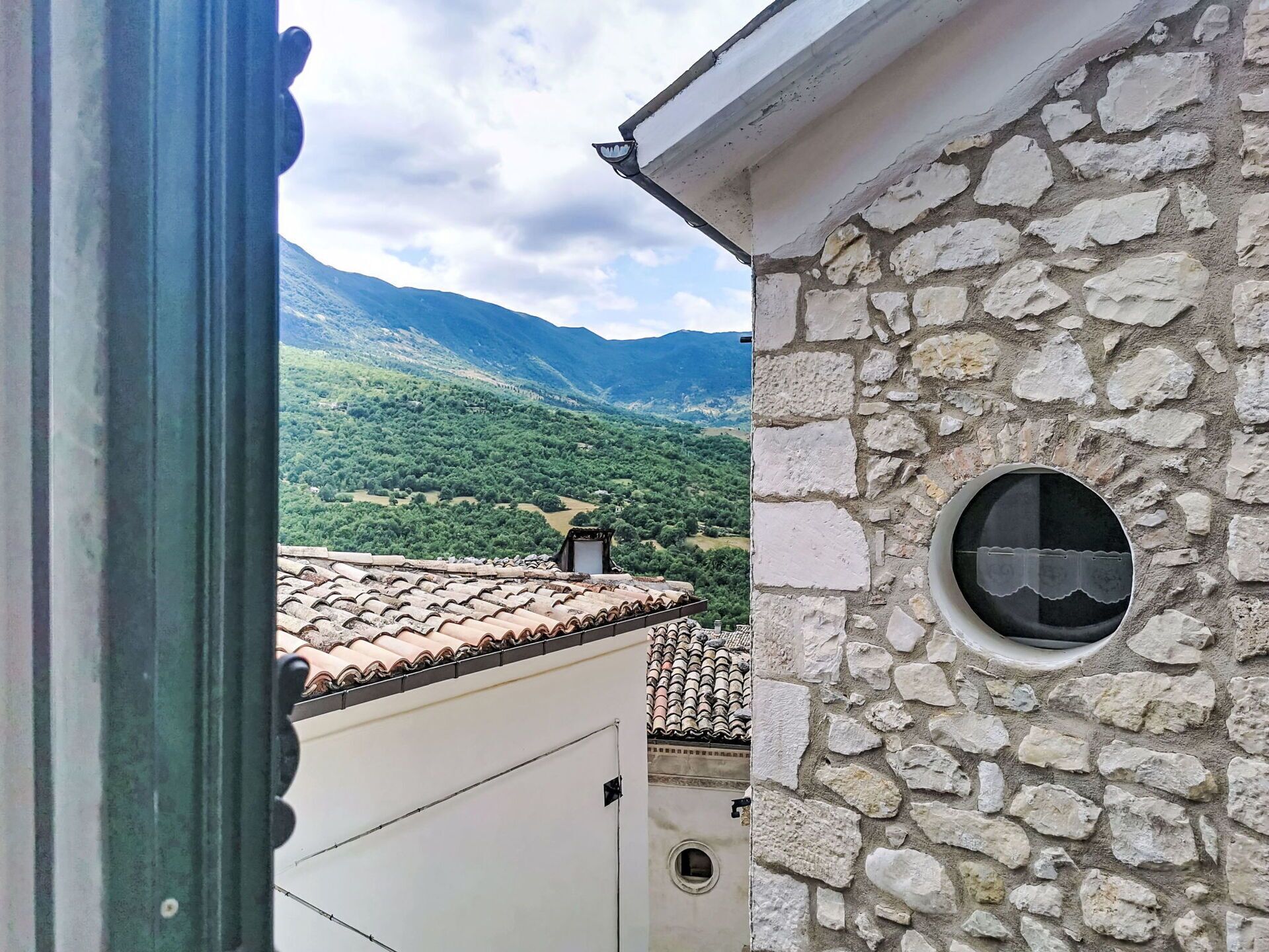 A stone building with a tiled roof, a circular window, and a view of green hills.