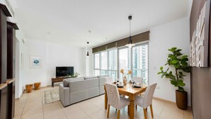A modern living room with a grey sofa, dining table with chairs, a TV, and a large window with a city view. - Cozy 1BR Steps From Burj Khalifa Mall (Dubai)
