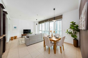 A modern living room with a grey sofa, dining table with chairs, a TV, and a large window with a city view. - Cozy 1BR Steps From Burj Khalifa Mall (Dubai)