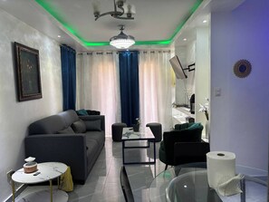 Ein modernes Wohnzimmer mit einem grauen Sofa, einem Glastisch und einem schwarzen Sessel. Auf einem kleinen Tisch stehen eine Vase und eine Fernbedienung. Der Raum verfügt über einen Deckenventilator, eine Kristallleuchter und ein großes Fenster mit Vorhängen.