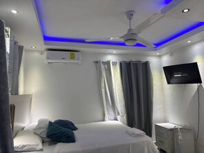 Ein Schlafzimmer mit einem Bett, einem Deckenventilator, einer Klimaanlage, einem Fernseher und Vorhängen.
