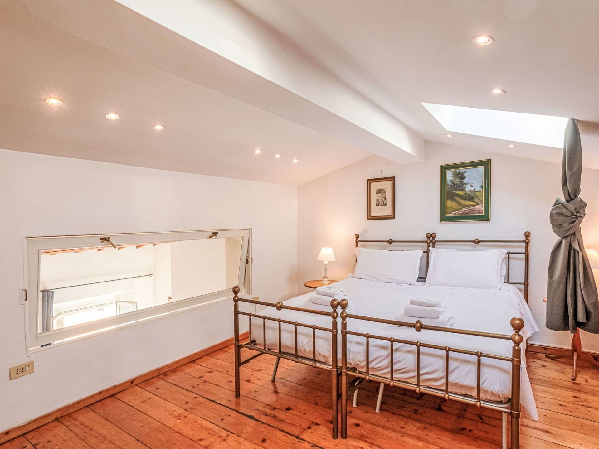 A bedroom with a skylight, a bed with white linens, a bedside table with a lamp, and a framed picture on the wall.