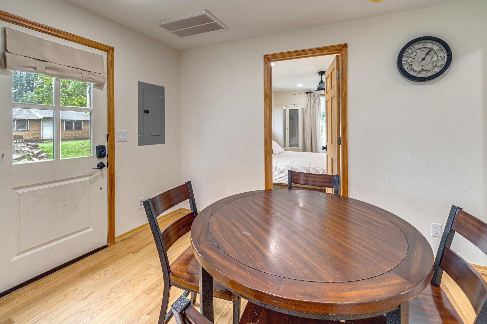 Un comedor con una mesa redonda de madera y cuatro sillas, un reloj en la pared y una puerta que da a otra habitación.